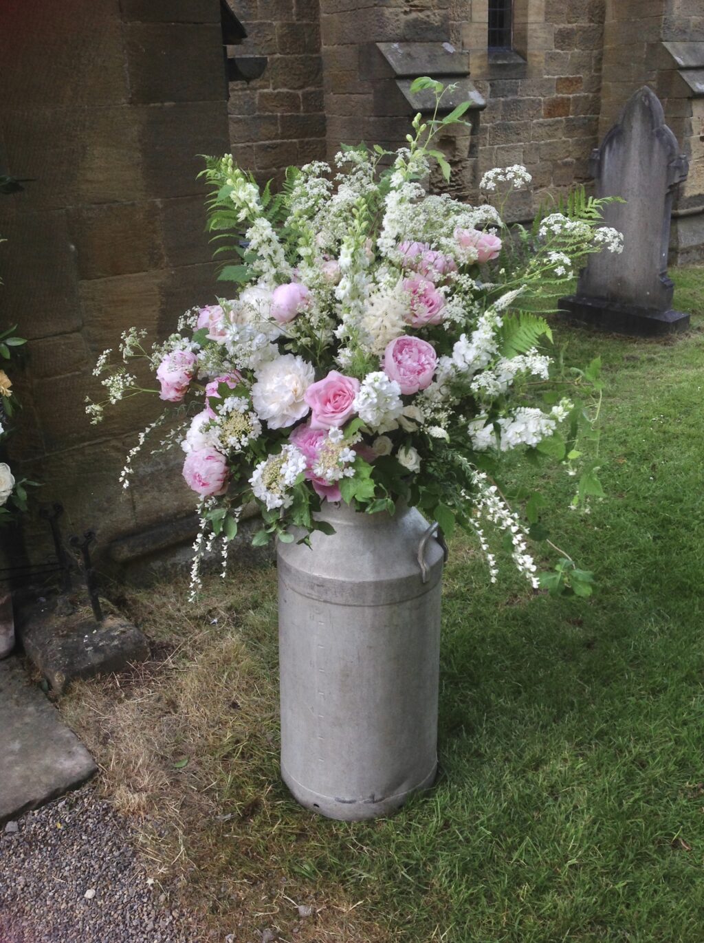 Wedding flowers - a milk churn outside the church is packed with scents from pink roses, white peonies and stocks, and overflows with frothy white cow parsley and ammi for a wildflower style church wedding