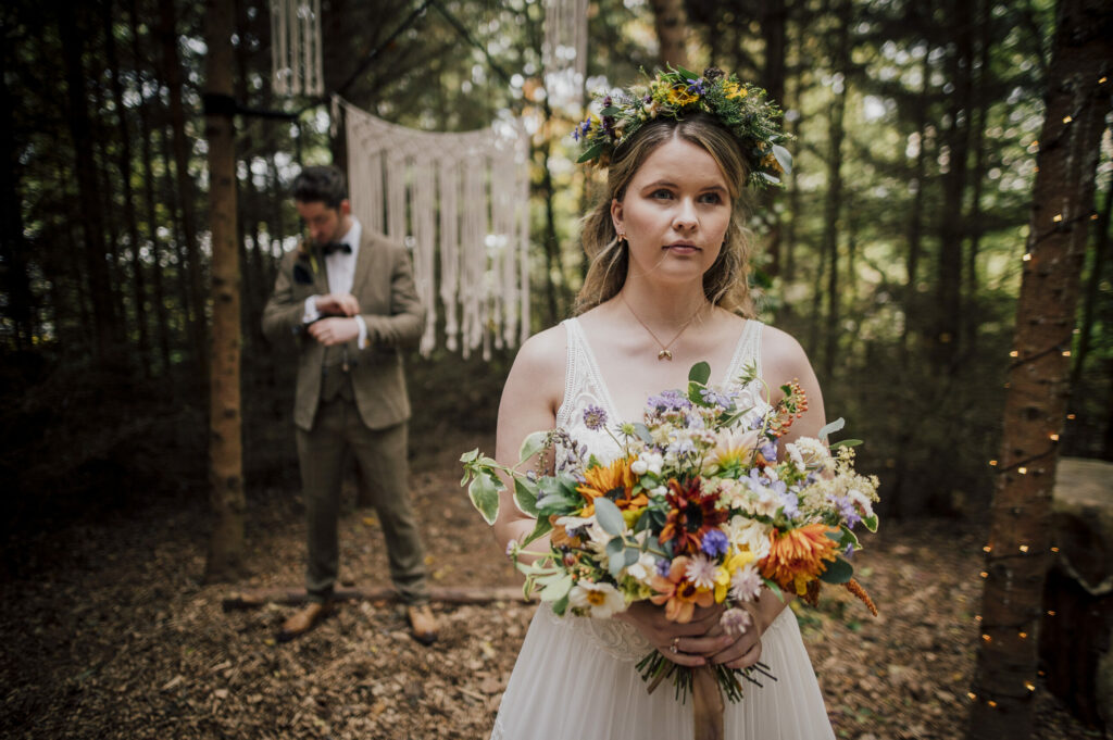 A bride wearing a wildflower flower crown for a woodland wedding holds a bouquet of vibrant summer flowers. Camomile and Cornflowers. Photo Dearest Love Photography.