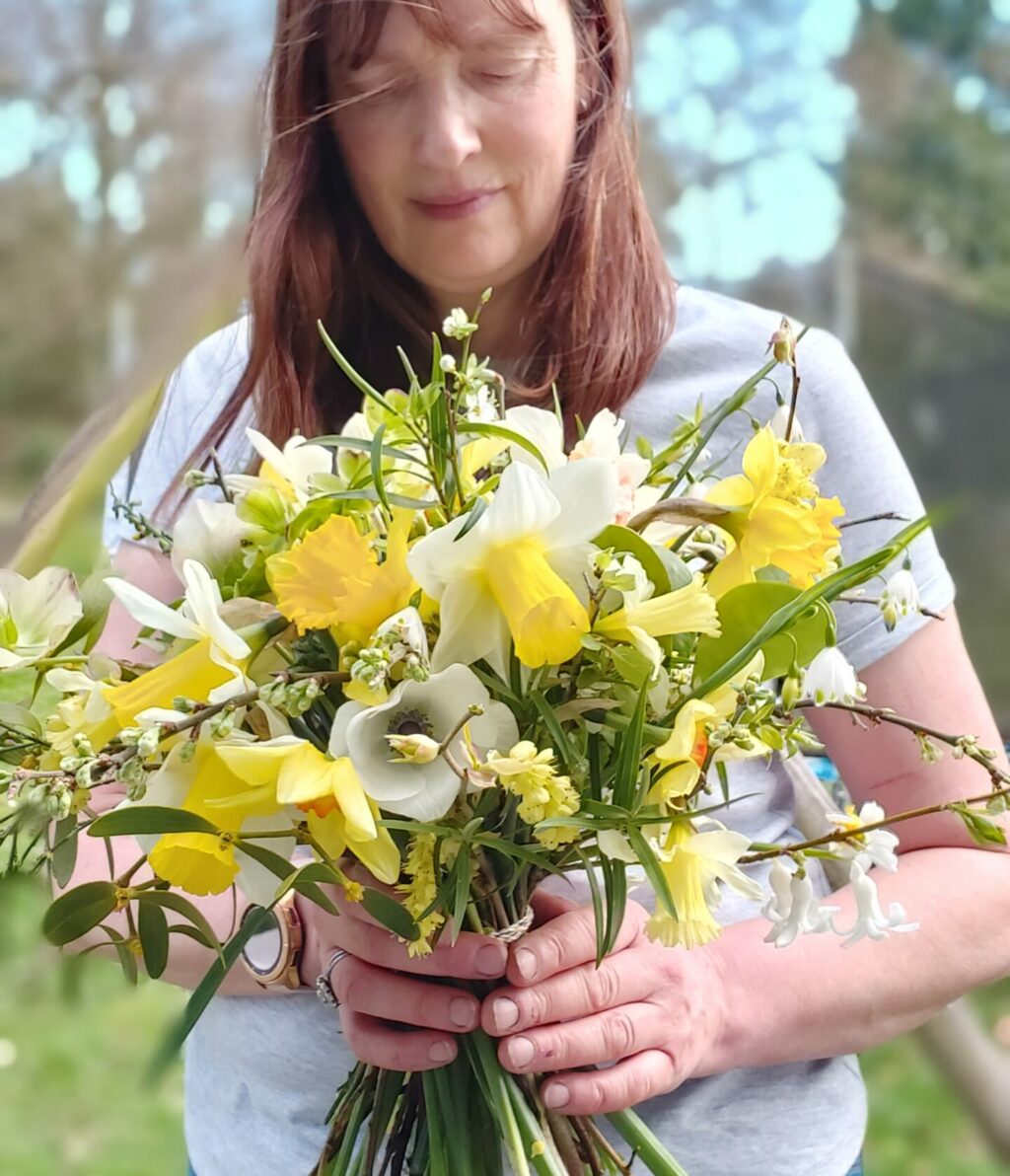 Kate of Camomile and Cornflowers stands in her flower field against a blue sky holding a bouquet of yellow daffodils.