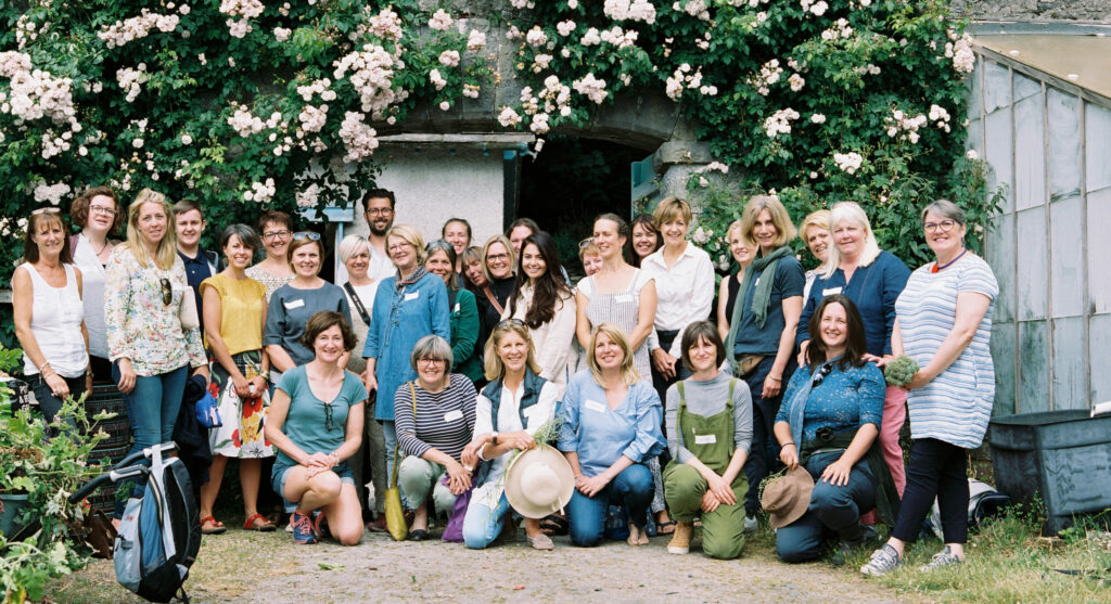 Members of the Flowers from the Farm North West regional group gather togther for a meeting and smile for a happy group photo in front of doorway in a beautiful stone wall clad with a blush pink rambling rose.