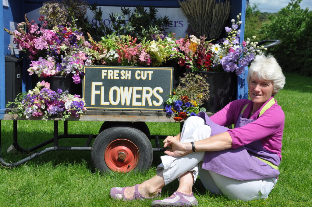 Gill Hodgson, founder of Flowers from the Farm, sits on the grass in front of a blue trolley filled with buckets of freshly picked British cut flowers from her farm.