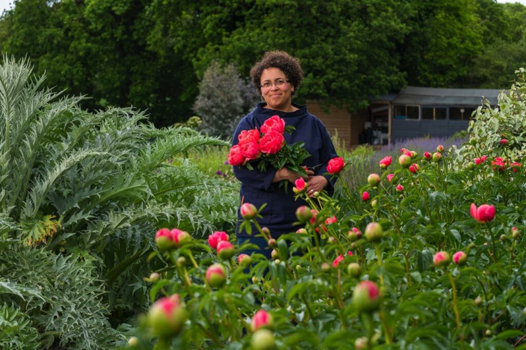 Cel of Forever Green Flower Company on her Norfolk flower farm in June at the height of peony season. She stands next to a bed of bright pink peonies just about to break bud, and holds a bunch of freshly cut, fully open peonies in her hands.