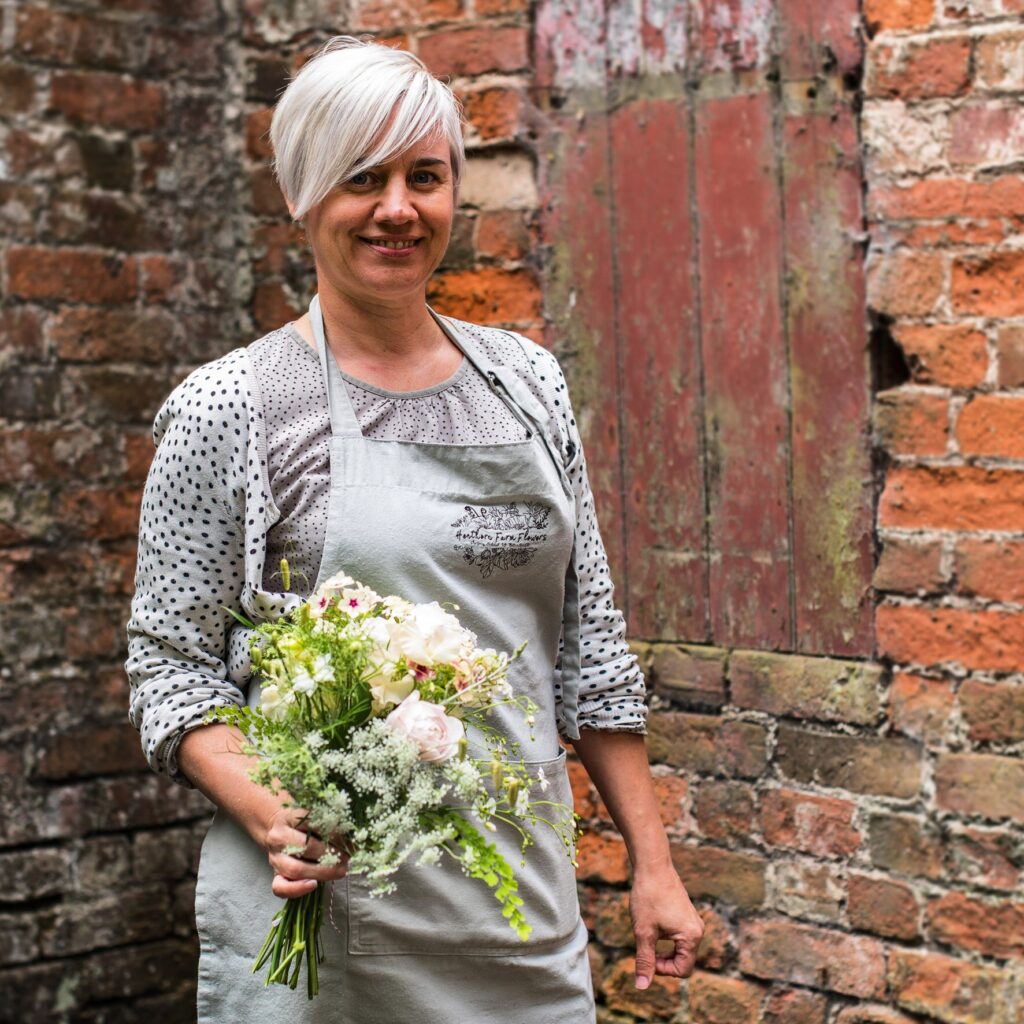 Kirsten of Henthorn Farm Flowers holds a seasonal boquuet of delicate British cut flowers - frothy Queen Anne's Lace, love-in-a-mist, snapdragons and phlox Creme Brulee. She stands against a rough brick terracotta wall as a backdrop.