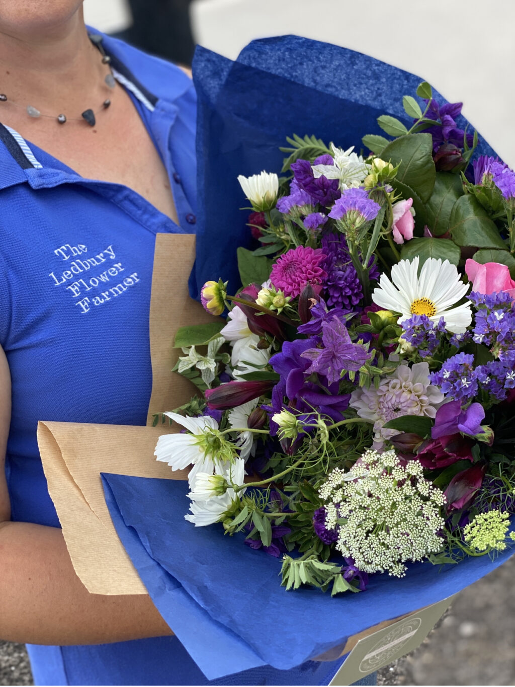 Rozanne, The Ledbury Flower Farmer, holds a bouquet of bright late summer flowers in blues, pinks and whites wrapped in vibrant blue tissue.
