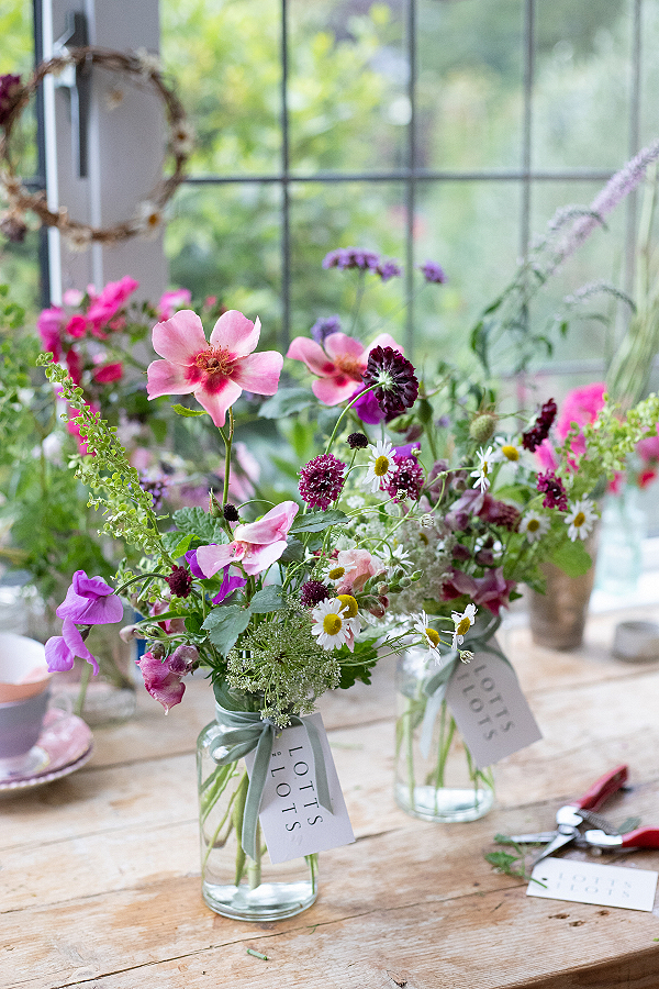 Jam jars hold brightly coloured summer blooms of garden gathered flowers. They're on a wooden table in front of a window which looks out onto the garden where they grew.
