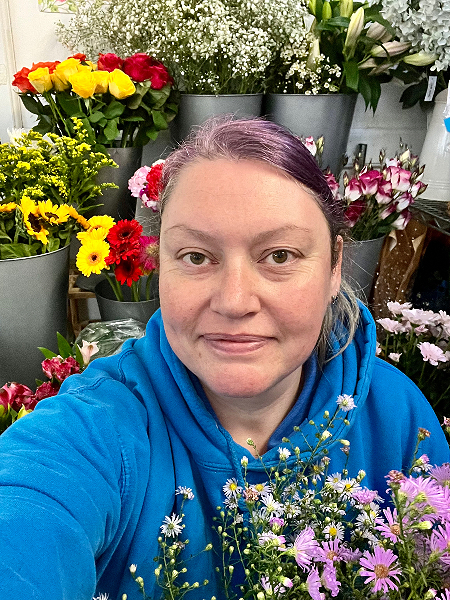 Sarah Francis of The Flower Academy, Derbyshire, in her workshop, holding a bunch of asters with colourful red and yellow flowers in the background.