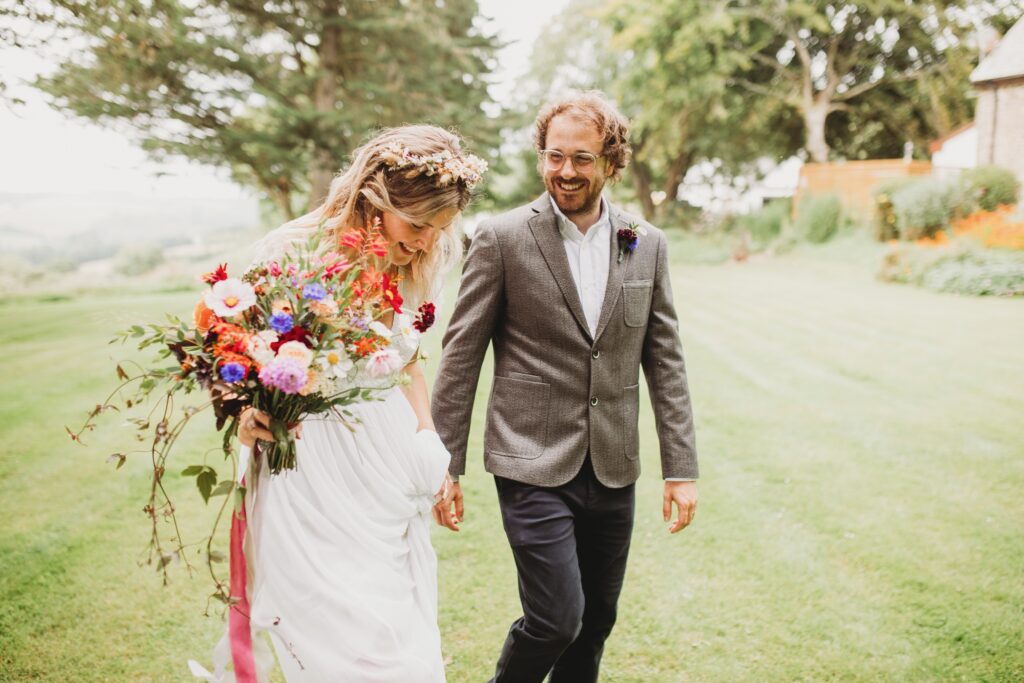 A colourful summer wedding bouquet is carried by the bride as she walks through the garden with her new husband. Flowers by Sweet Peas and Sunflowers. Photo: Holly Collings Photography.