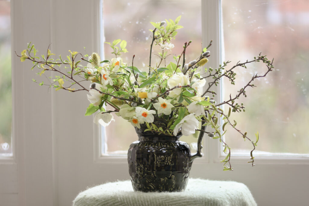 A dark green ceramic jug packed with seasonal spring flowers: narcissi, hellebores, pussy willow and bursting buds of bright green foliage.Tuckshop Flowers