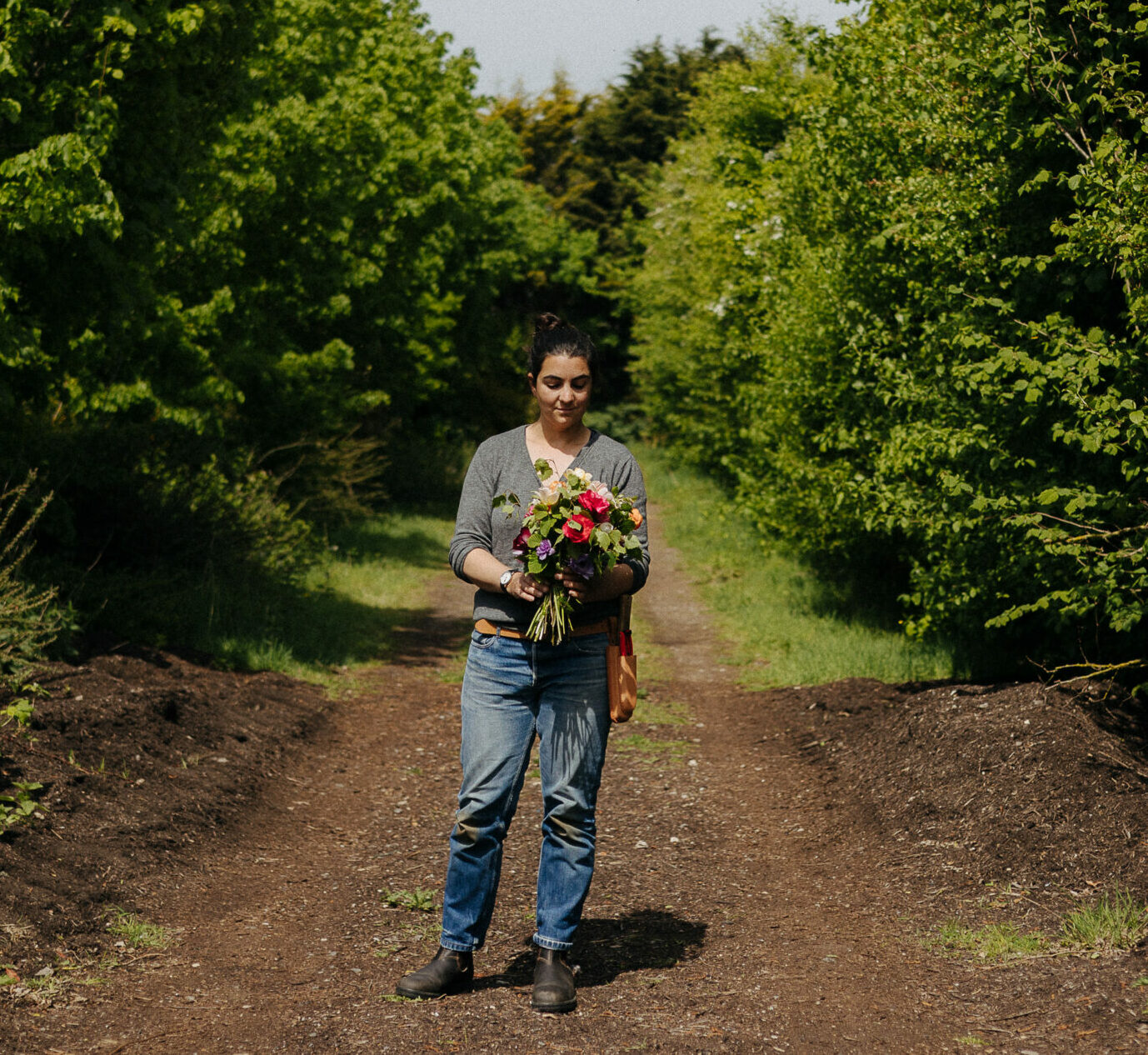 Susie Ross of Wivenhoe Flowers stands with a hand tied bouquet of her flowers on a quiet woodland path