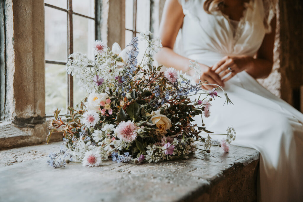 A wildflower style bride's bouquet rests on a windowsill with the summer flowers catching the light as the bride sits nearby. Yewbarrow Farm Flowers.